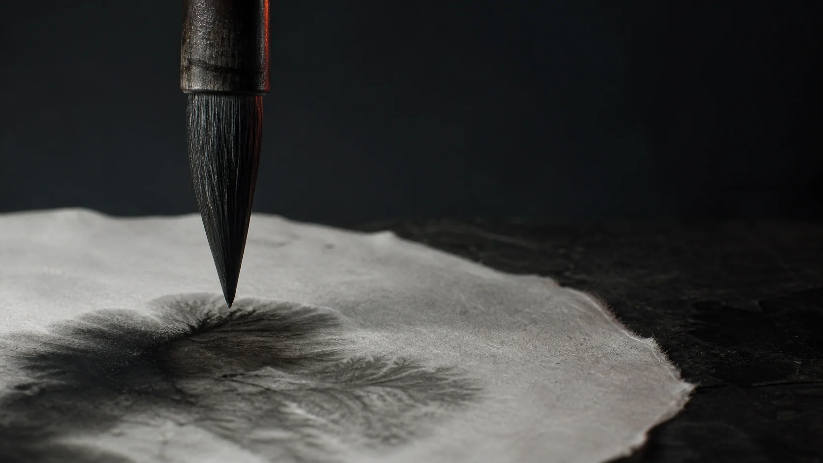 An ink brush suspended above fibrous paper as wet ink spreads across the surface